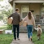 A mother and father walking down a tree-lined neighborhood sidewalk at golden hour, swinging their small child between them by the hands
